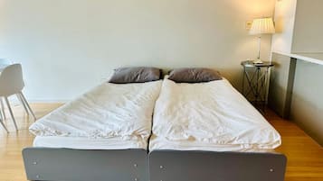 A double bed with white bedding and two pillows, a small bedside table with a lamp, and a chair in a room with wooden flooring and white walls.
