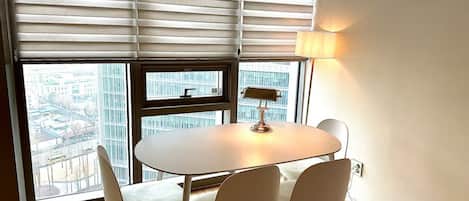 Une pièce moderne avec une table ronde, quatre chaises, une lampe et de grandes fenêtres équipées de stores.