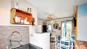 Une cuisine compacte équipée d’un évier, d’un four à micro-ondes et d’une étagère en bois située au-dessus. On y trouve également un coin repas avec une table et des chaises, ainsi qu’une fenêtre avec des rideaux.