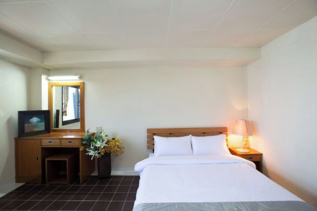 Una camera d'albergo con un letto, un comodino, una lampada, una scrivania con uno specchio e un piccolo armadietto.