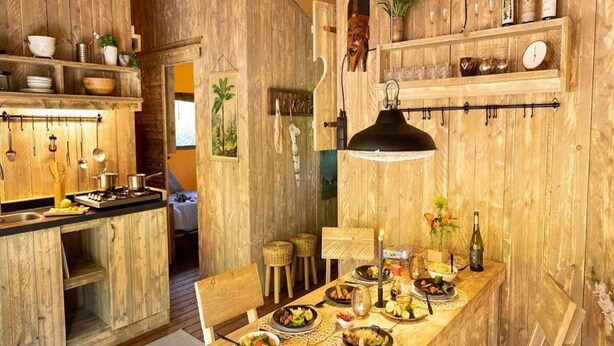 A rustic wooden kitchen with a dining area, featuring a long wooden table set for a meal, a hanging light fixture, and various kitchen utensils and appliances. - Luxury Sunshine Tent in Dochamps (Manhay)