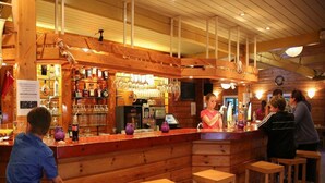 A rustic wooden bar area with stools, a counter, and shelves stocked with bottles and glasses. - Luxury Sunshine Tent in Dochamps (Manhay)