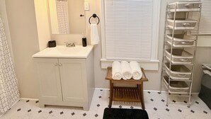 Ein Badezimmer mit einem weißen Waschbecken, einem Spiegel, einem Handtuchhalter, einem Fenster mit Jalousien und einem Wäscheständer mit weißen Handtüchern.
