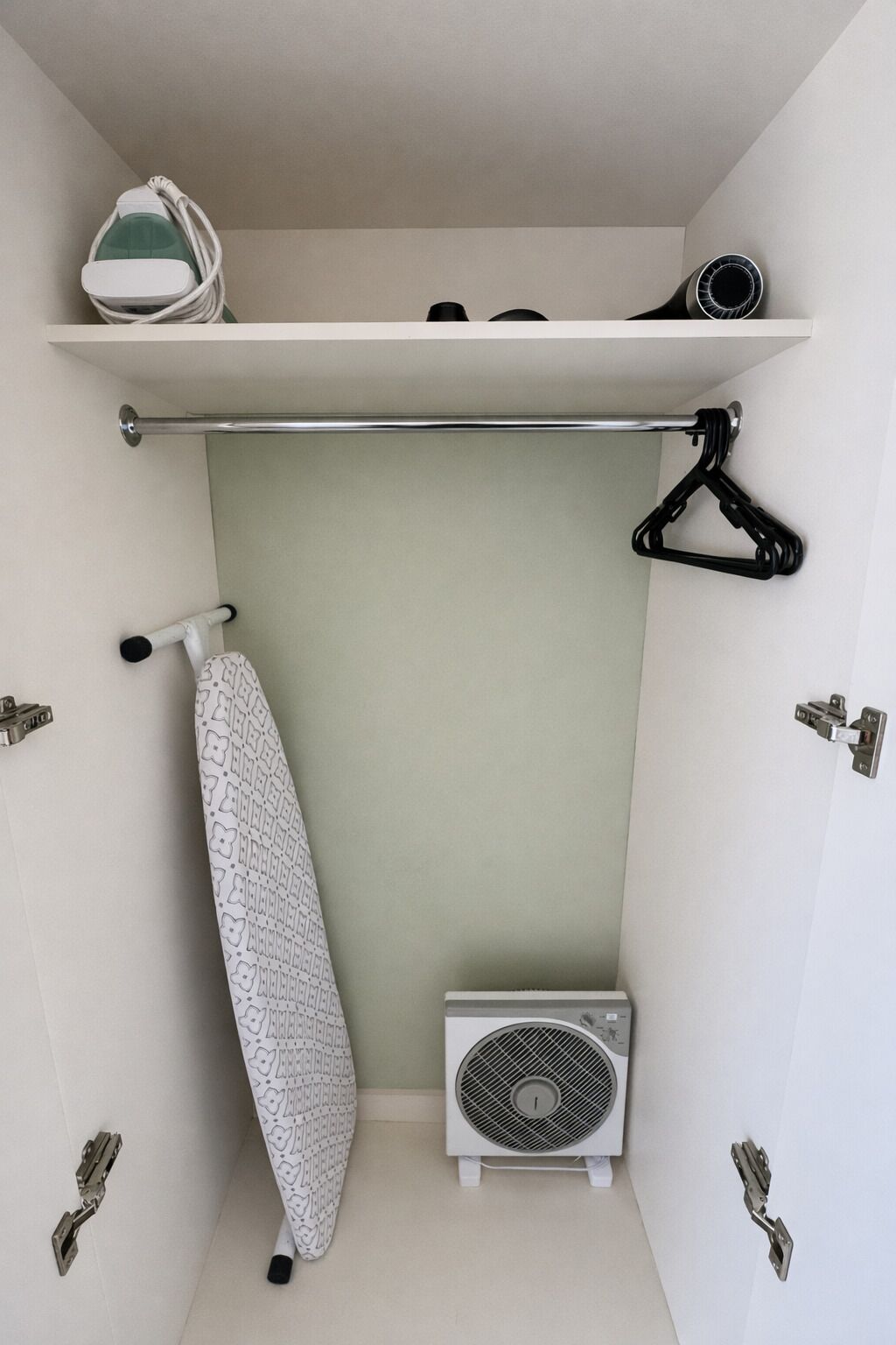A small room with a white ironing board, a hanging rack, a radiator, and a ceiling fan.