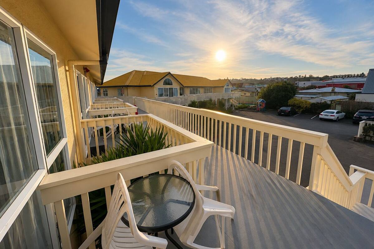 A balcony with a glass table and chairs, overlooking a parking area and buildings.