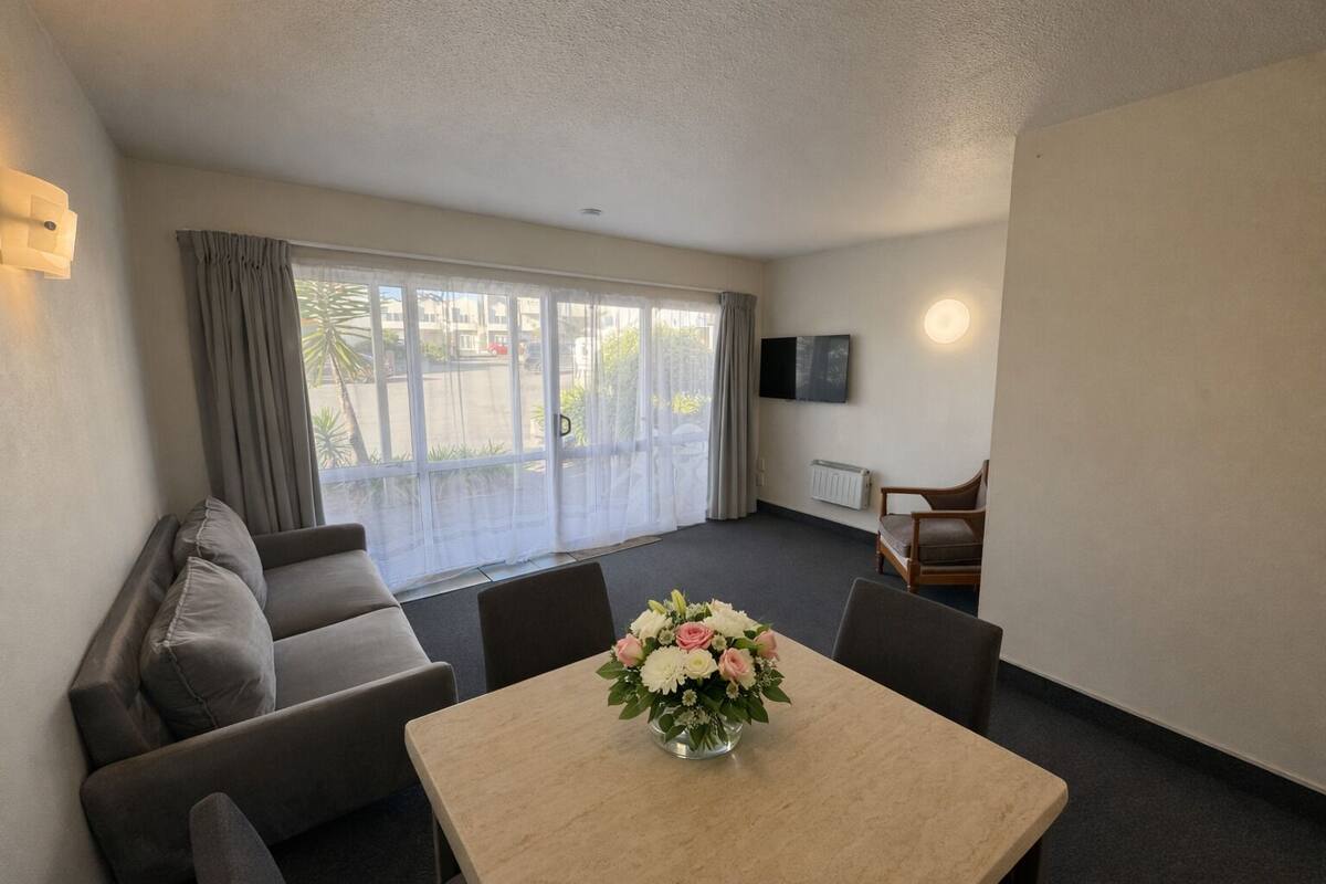 A room with a sofa, a table with a vase of flowers, a TV on the wall, and a sliding door to the outside.