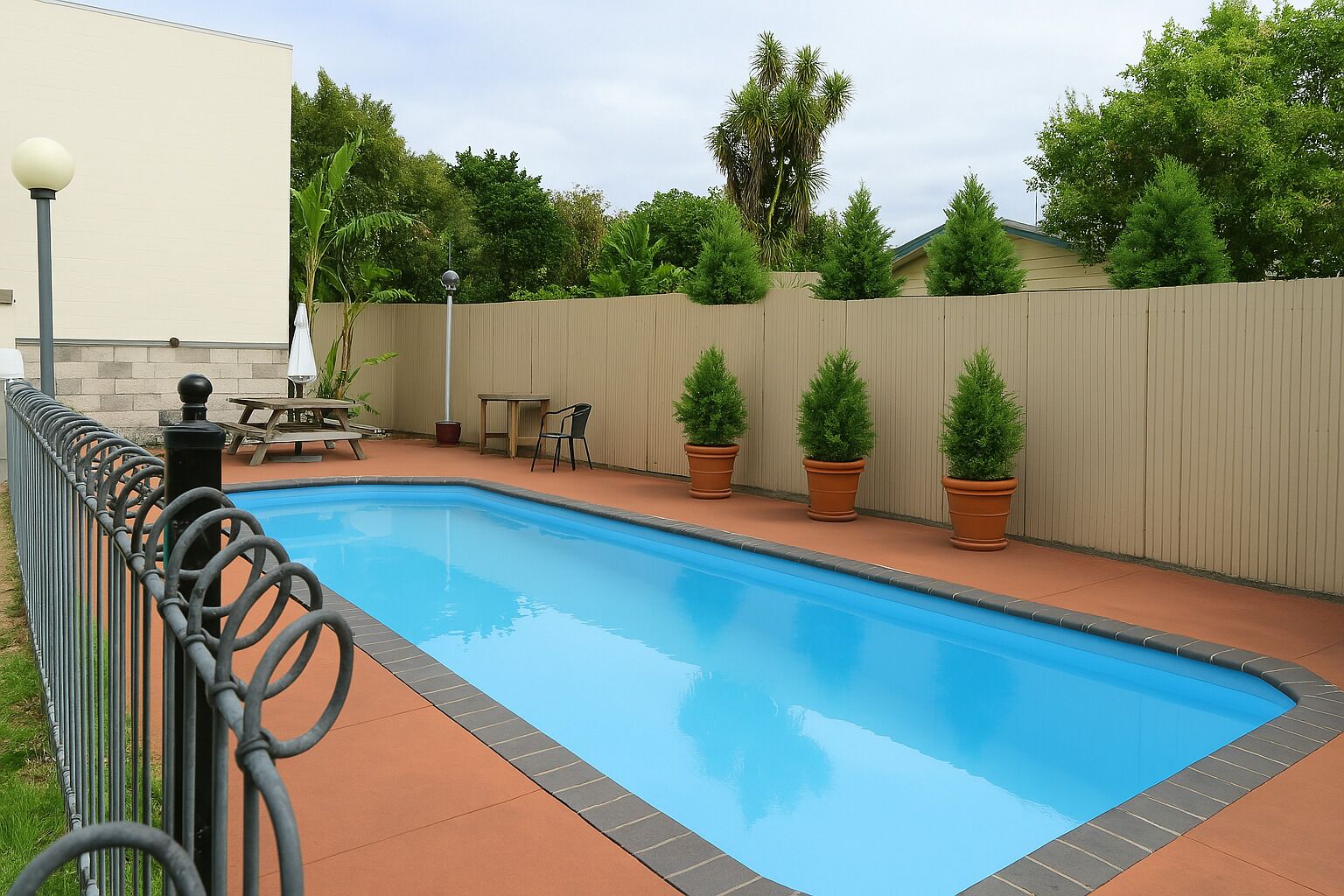 A rectangular swimming pool with a metal fence, surrounded by potted plants and a picnic table.