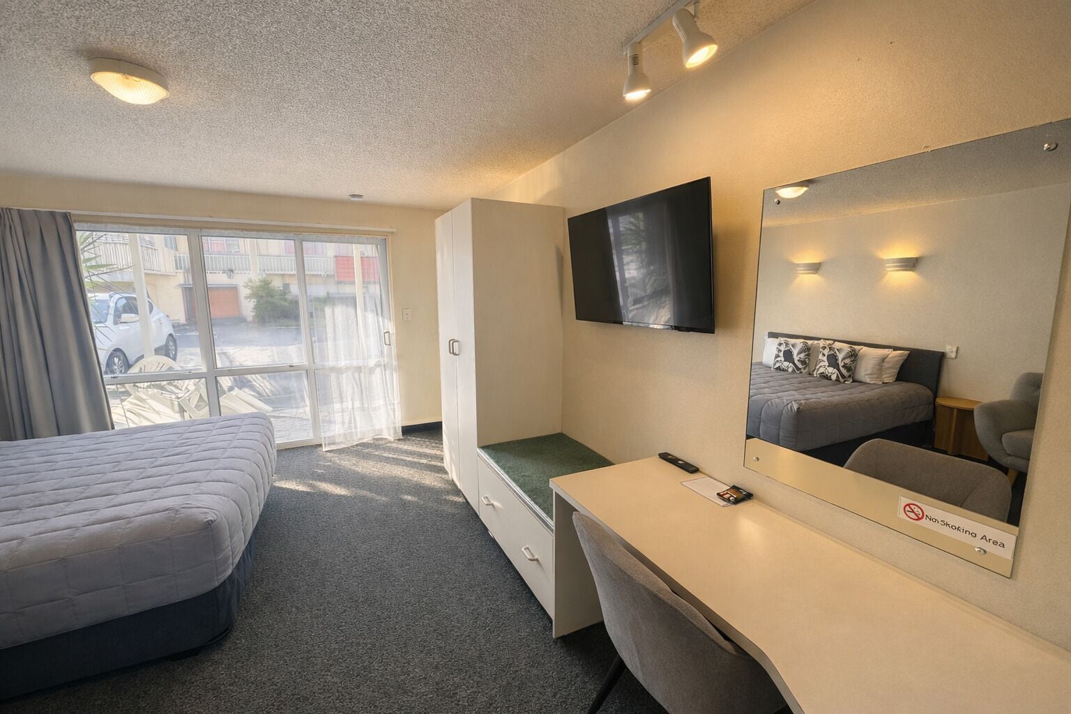 Une chambre d’hôtel équipée d’un lit, d’un bureau, d’une chaise, d’une télévision et d’une porte coulissante donnant sur l’extérieur.