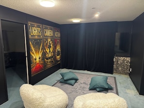 A room with a dark wall, a poster with lights, camera, and action, a flat-screen TV, and two fluffy chairs. - BK's Fountain Court Motel (Napier)