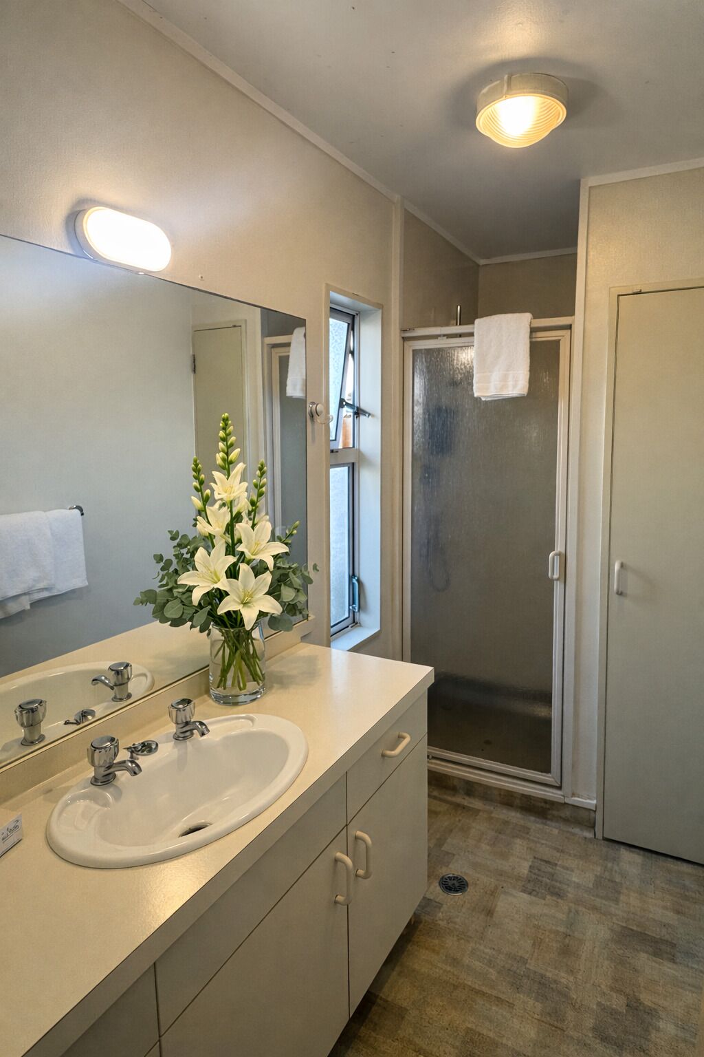 Une salle de bain avec un grand miroir, un lavabo, un vase de fleurs, une douche et une porte.