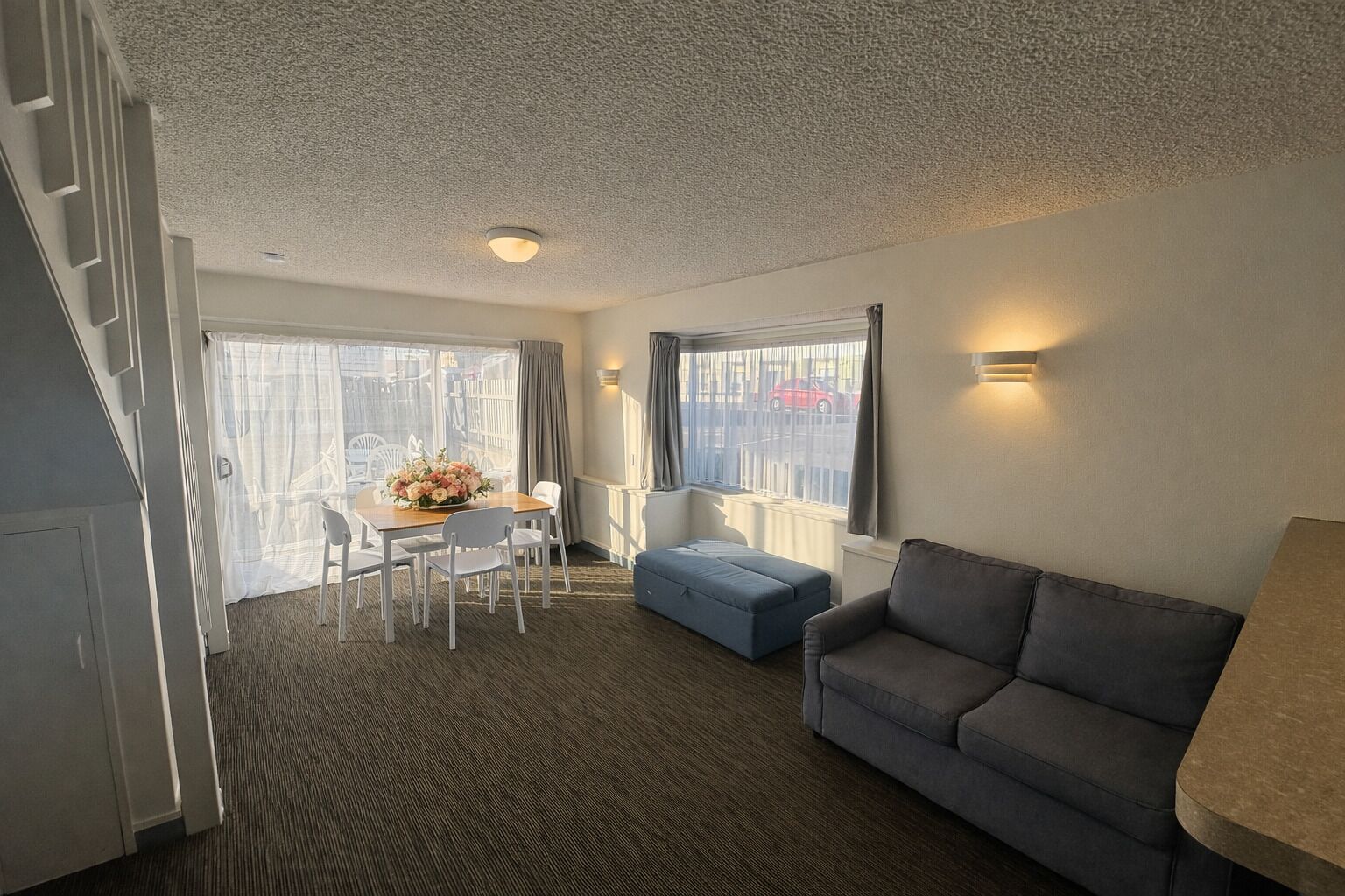 Habitación de hotel con una mesa de comedor, sillas, un sofá, un ventanal con cortinas e iluminación en el techo.