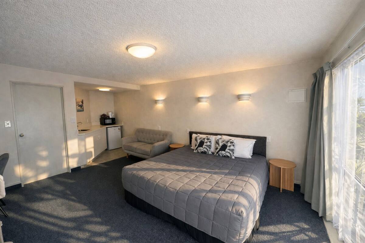 A hotel room with a bed, a chair, a small table, a kitchenette, and a window with curtains.