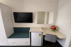 Quarto de hotel compacto com cama, pequena escrivaninha, geladeira, televisão, espelho e um vaso de flores.
