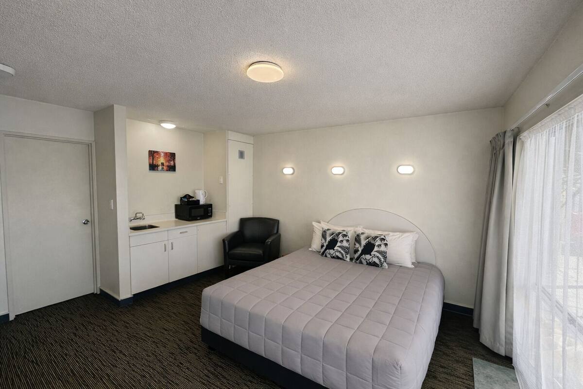 A hotel room with a bed, a chair, a desk with a microwave, and a window with curtains.