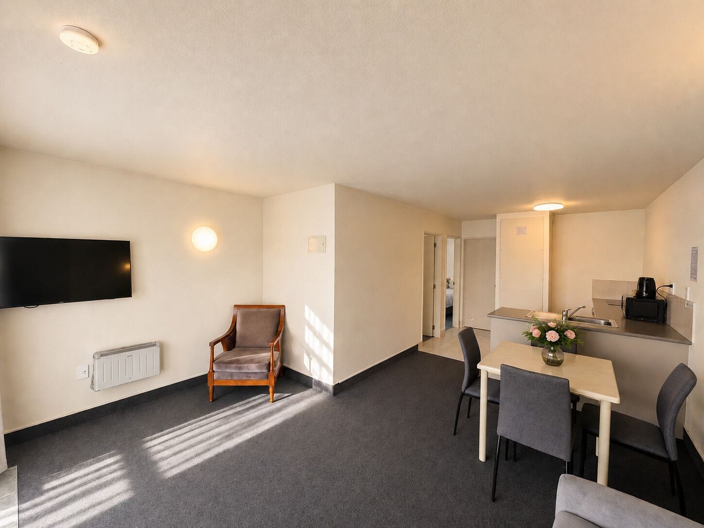 A hotel room with a TV, a chair, a dining table with chairs, a small kitchen area, and a radiator.