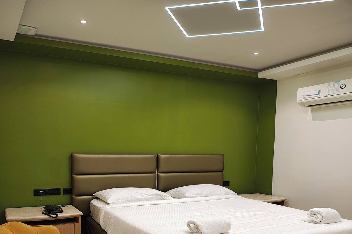 A hotel room with a bed, a bedside table with a phone, a chair, and a wall-mounted air conditioner.