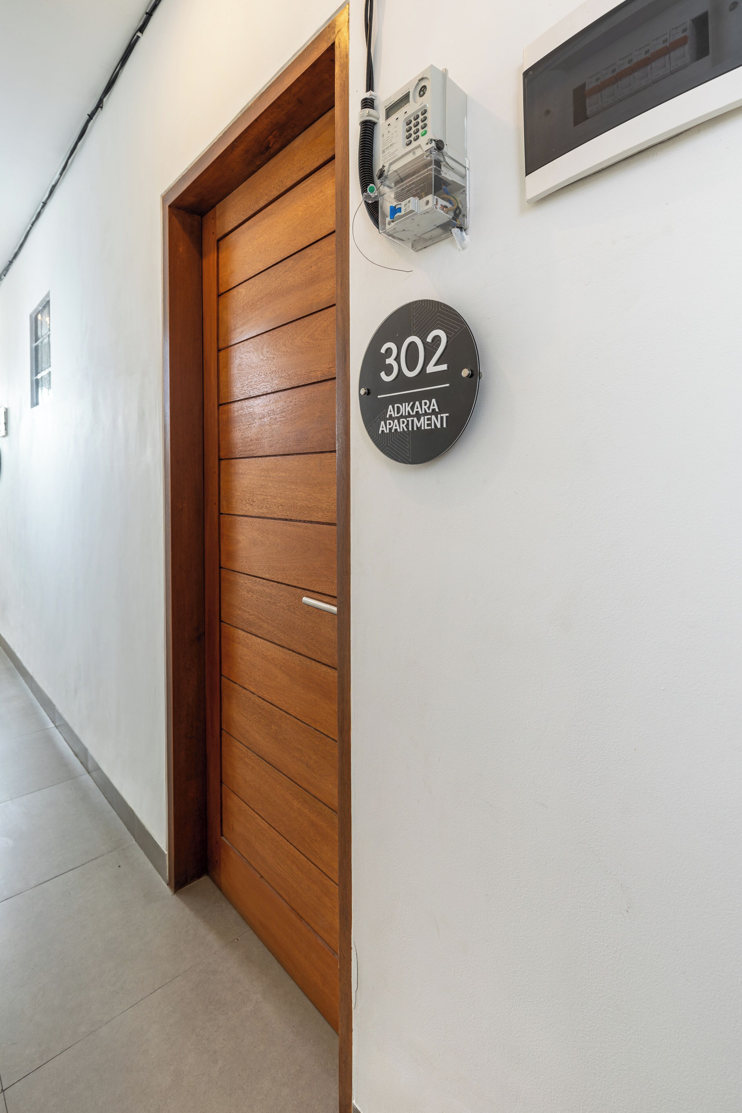 Un couloir avec une porte en bois, un interphone fixé au mur et une pancarte indiquant « Appartement 302, immeuble Adikara ».