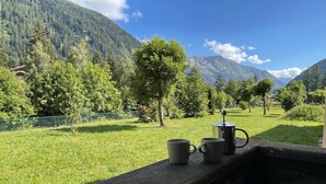 A wooden bench with two mugs and a coffee pot, overlooking a grassy field with trees and mountains in the background.