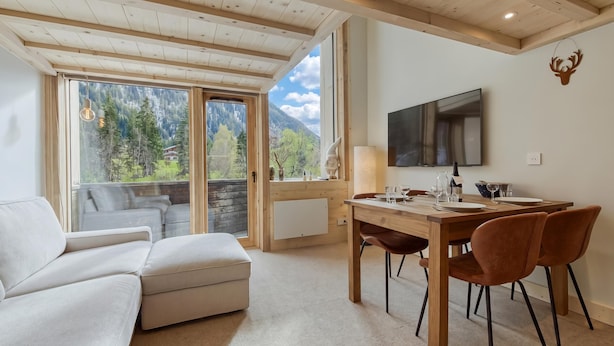 A modern living room with a sofa, dining table, and chairs, large windows with a mountain view, and a mounted television.
