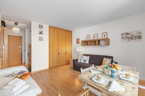 A room with a bed, a sofa, a dining table with plates and cutlery, a wooden wardrobe, and a wall with framed pictures.