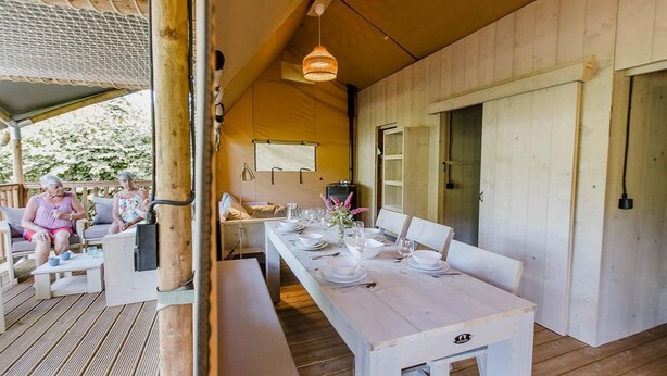 A wooden deck with a dining table set for four, a bench, and a covered seating area with two people.
