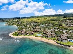 An aerial view of a coastal resort with multiple bungalow-style buildings, a central pool, and a beach.