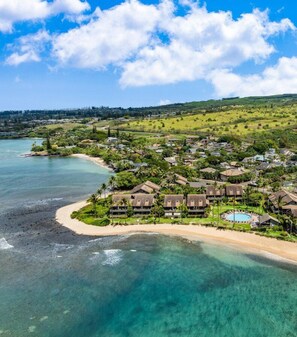 Vue aérienne d’un complexe hôtelier situé sur la côte, avec plusieurs bâtiments de style bungalow, une piscine centrale et une plage.