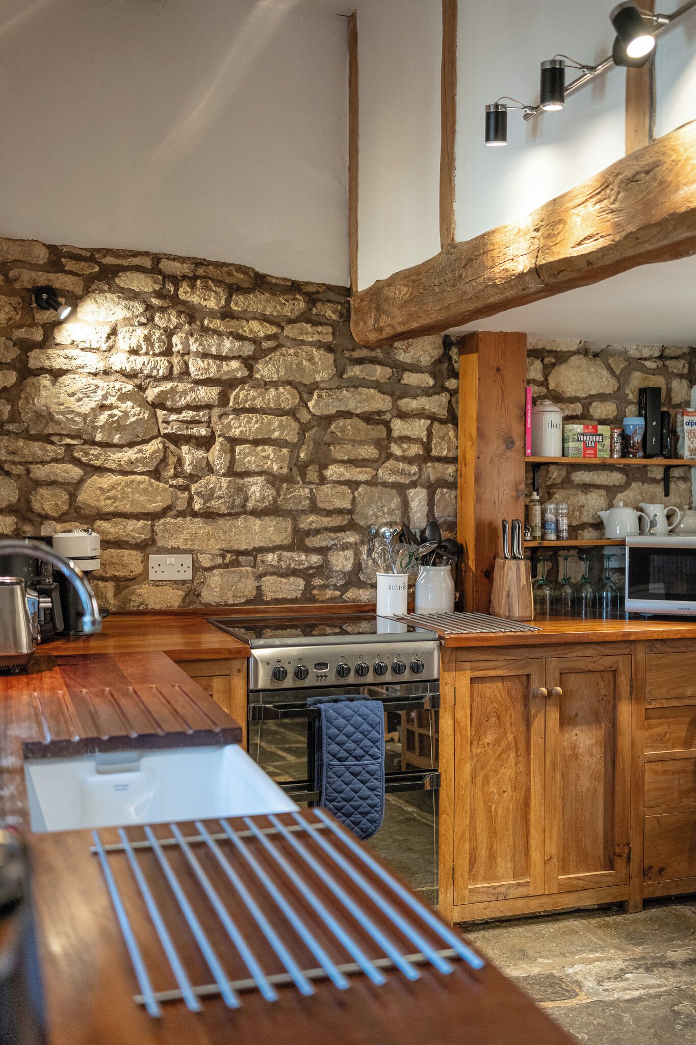 A kitchen with a stone wall, wooden beams, a stove, and various kitchen utensils.