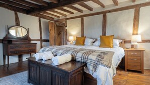 Una camera da letto con travi a vista in legno, un letto grande con coperta a quadri, una cassettiera in legno, un comodino con una lampada e uno specchio sulla cassettiera.
