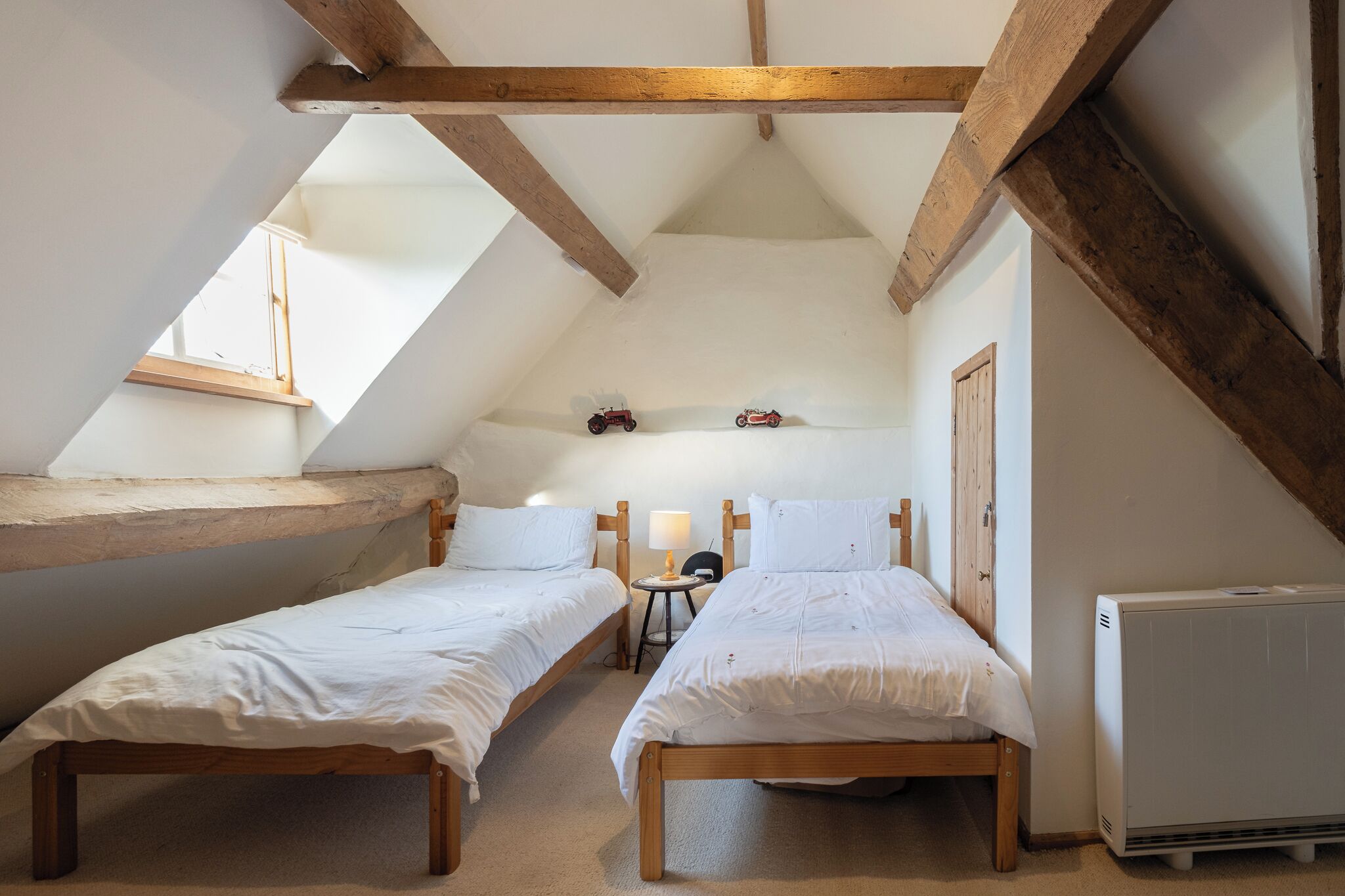 A room with two wooden beds, a small table with a lamp, a radiator, and a sloped ceiling with wooden beams.