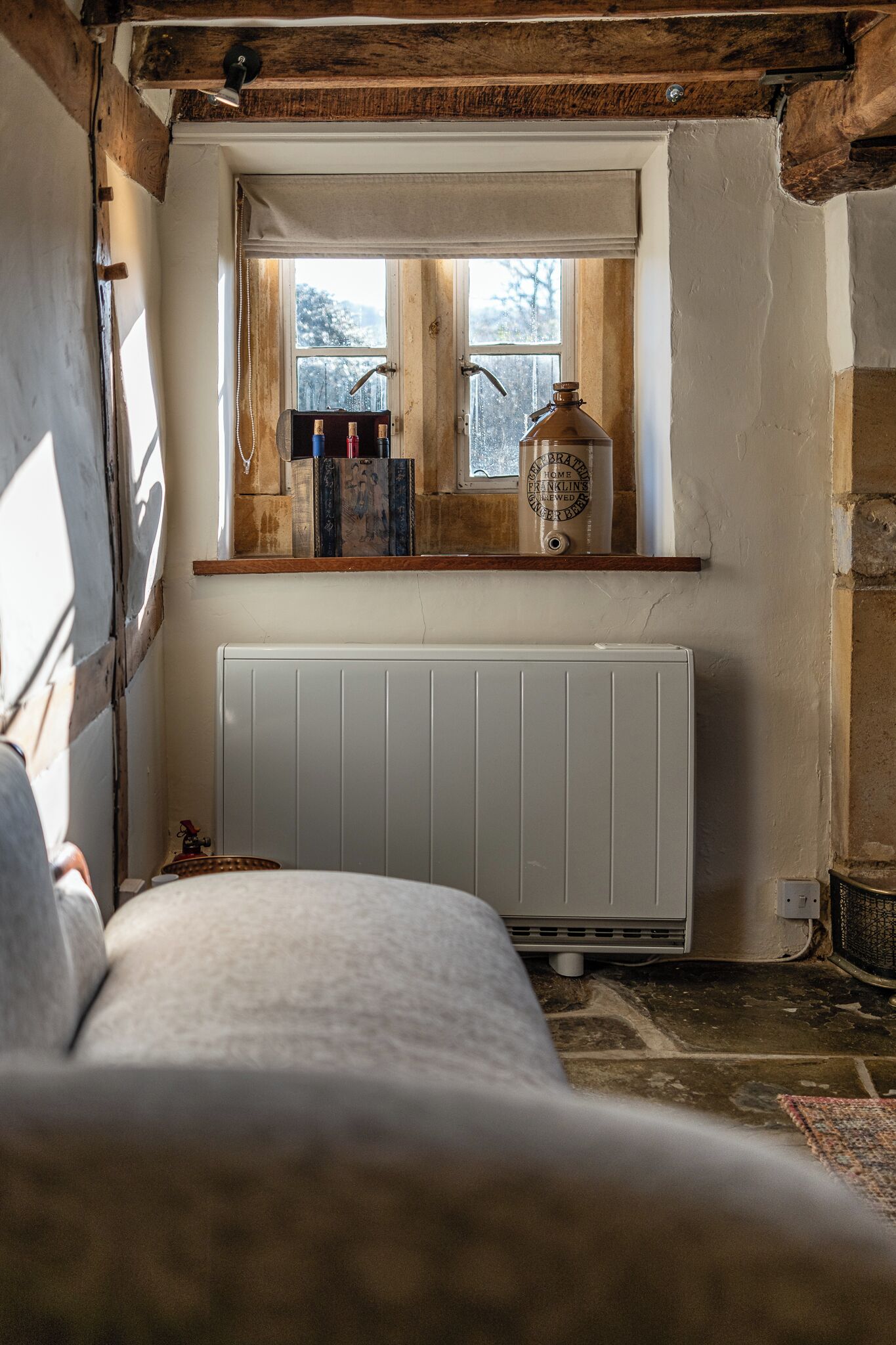A rustic room with a wooden beam ceiling, a window with bottles on the sill, a radiator below, and a bed in the foreground.