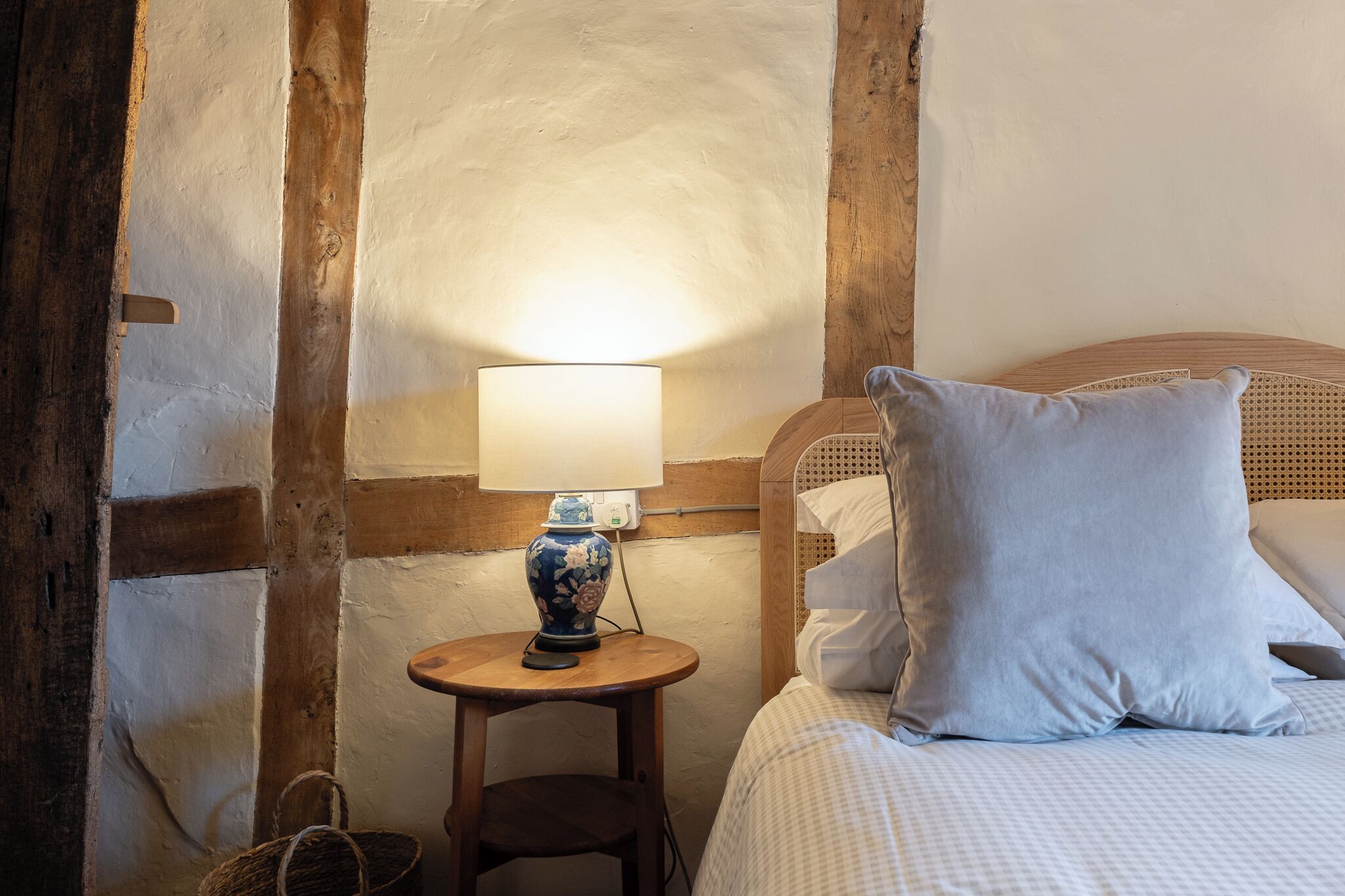 A bedroom with a wooden headboard, a bedside table with a lamp, a wicker basket, and a checkered bedspread.