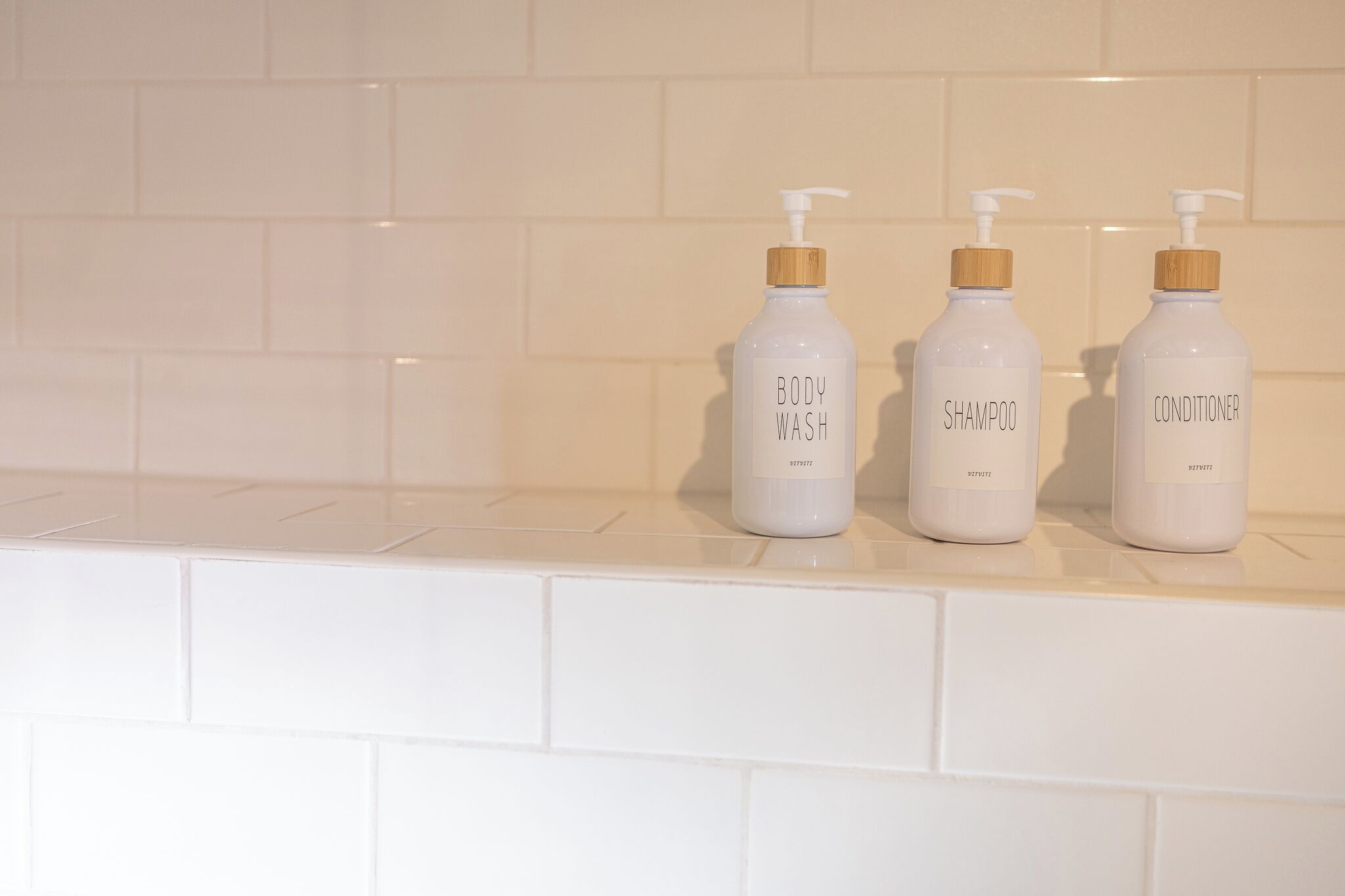 Three bottles of body wash, shampoo, and conditioner on a white tiled shelf.