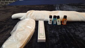 A neatly arranged set of toiletries on a bed with a dark blue cover, including a white towel, a box, and several bottles of personal care products. - Hotel High East (Papumpare)