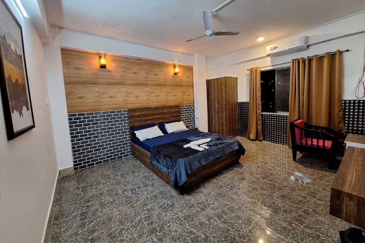 Ein Hotelzimmer mit einem Bett, einem Deckenventilator, einem hölzernen Kopfteil, einer schwarz-weißen Fliesenwand, einer Holztür und einem Fenster mit Vorhängen.