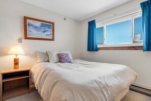 A bedroom with a bed, a nightstand, a lamp, a window with curtains, and a framed picture on the wall.