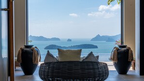A room with a large window overlooking a body of water and islands. There are two large vases with plants on either side of a wicker chair with cushions.