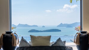 A room with a large window overlooking a calm sea and distant islands. There is a wicker chair with cushions and two decorative vases with tassels.