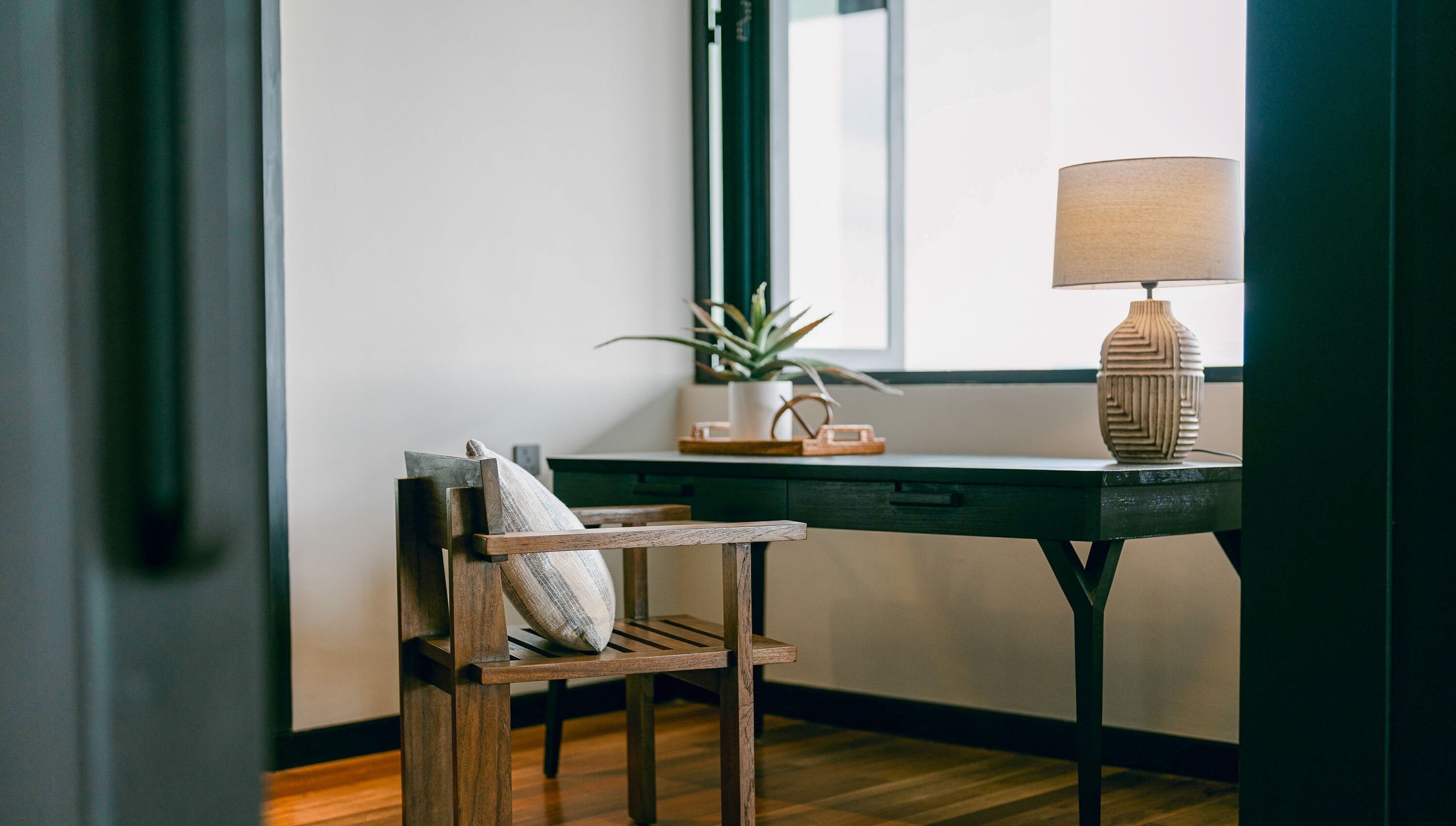 A wooden chair with a pillow, a desk with a lamp, and a potted plant.