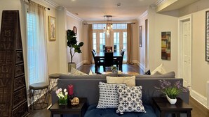 A living room with a dark blue sofa, a dining table with chairs, a large window with curtains, and a potted plant. - Ideal Group Getaway: Prime Location, Parking, Backyard, Sunroom & Pet-Friendly (Chicago)