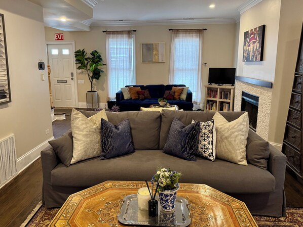 A living room with a brown sectional sofa, a coffee table with a tray, a blue and white vase, and a fireplace.