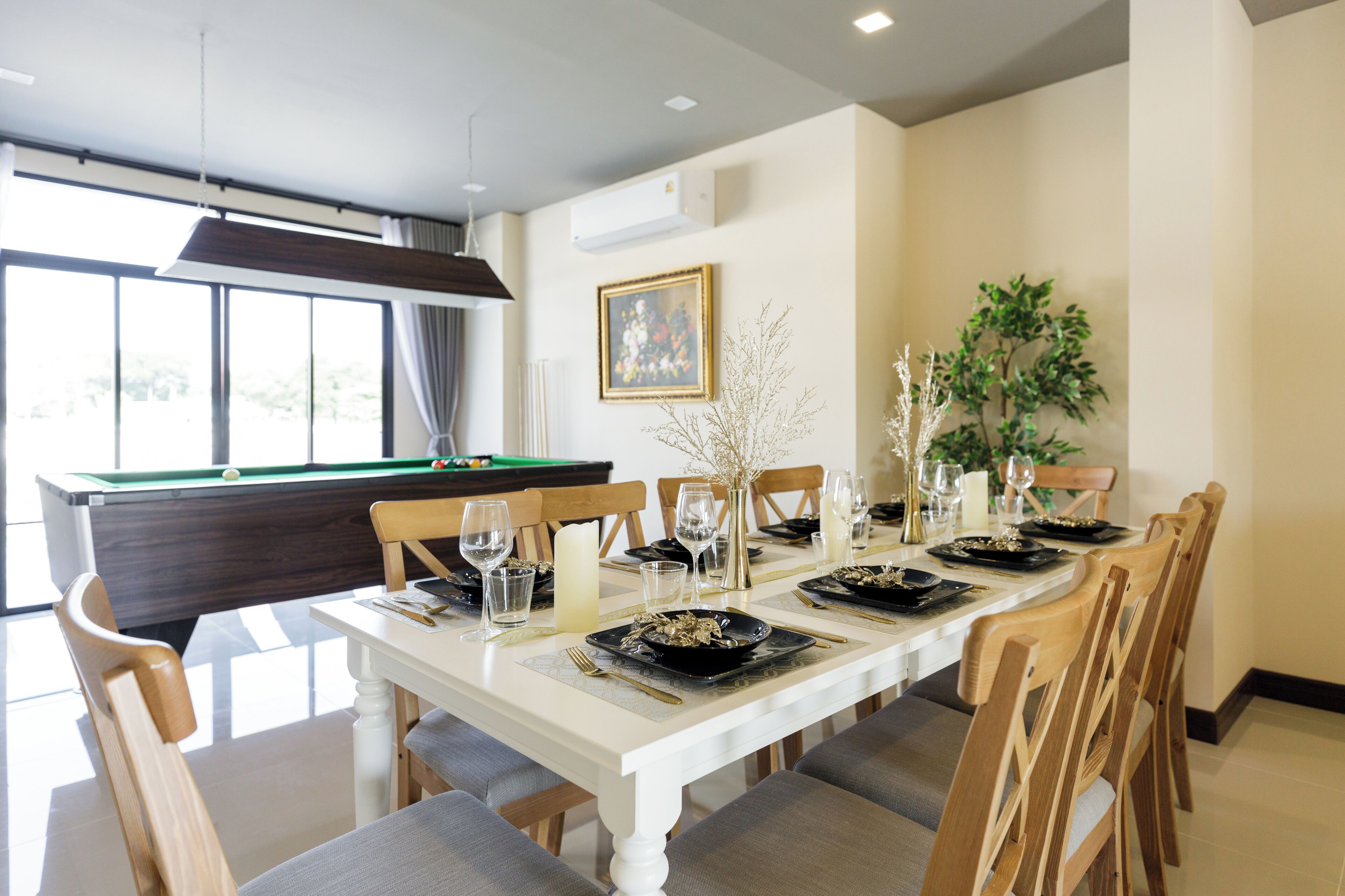 A dining area with a pool table, a dining table set for six with chairs, a vase with branches, and a framed picture on the wall.