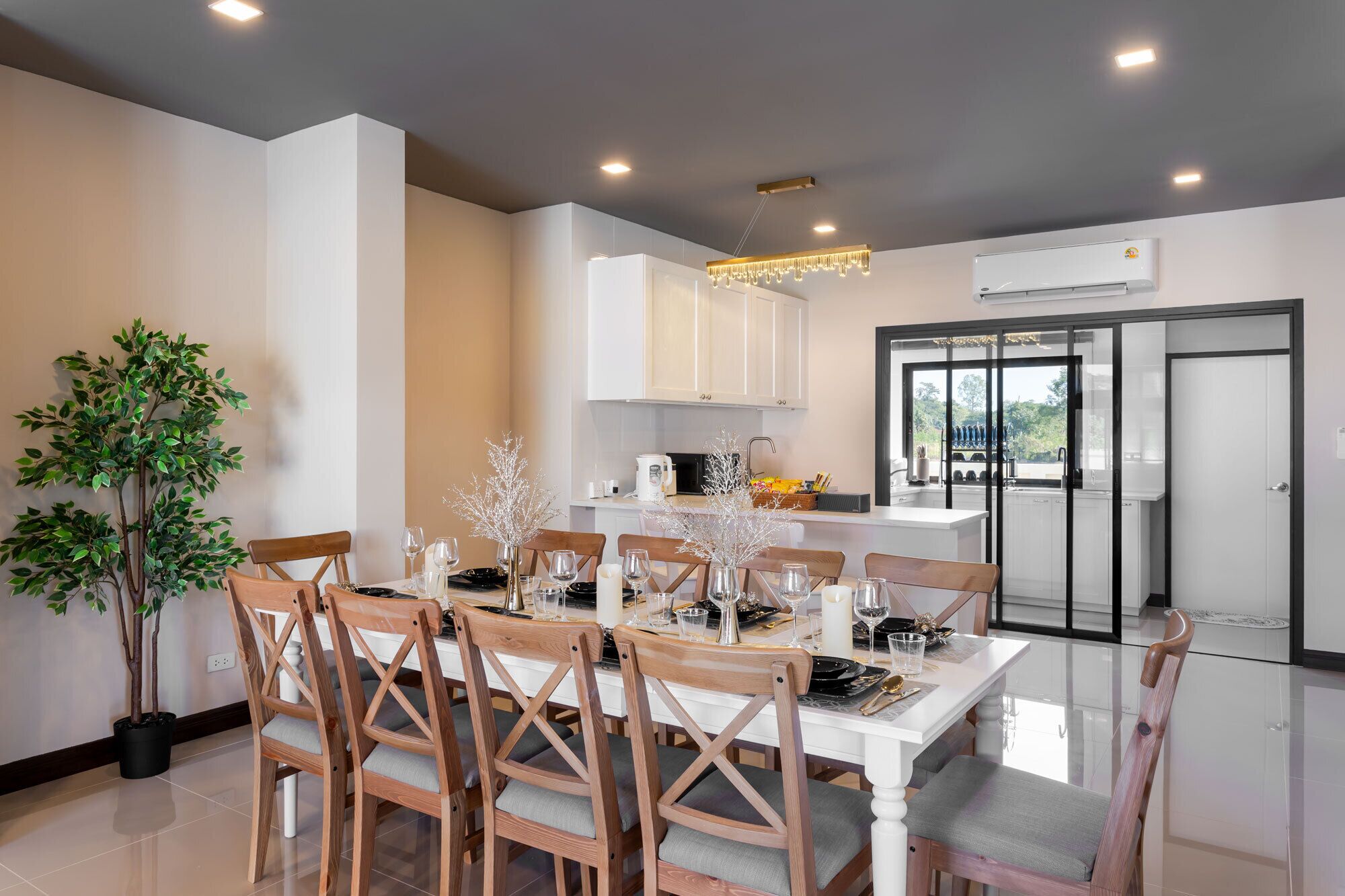 Un espace repas moderne avec une table blanche dressée pour six personnes, entourée de chaises en bois avec des coussins gris. Un pot de fleurs se trouve à gauche, et à l’arrière, on aperçoit un coin cuisine avec des meubles blancs et une porte à cadre noir.