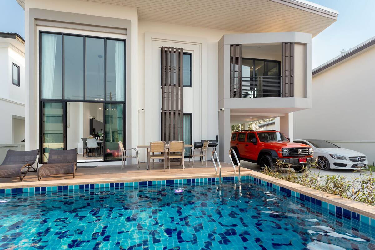 A modern two-story house with a swimming pool, outdoor seating, and a red SUV parked outside.