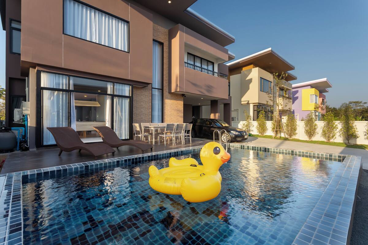 A modern pool with a yellow inflatable duck, a patio area with outdoor furniture, and a multi-story building with large windows.