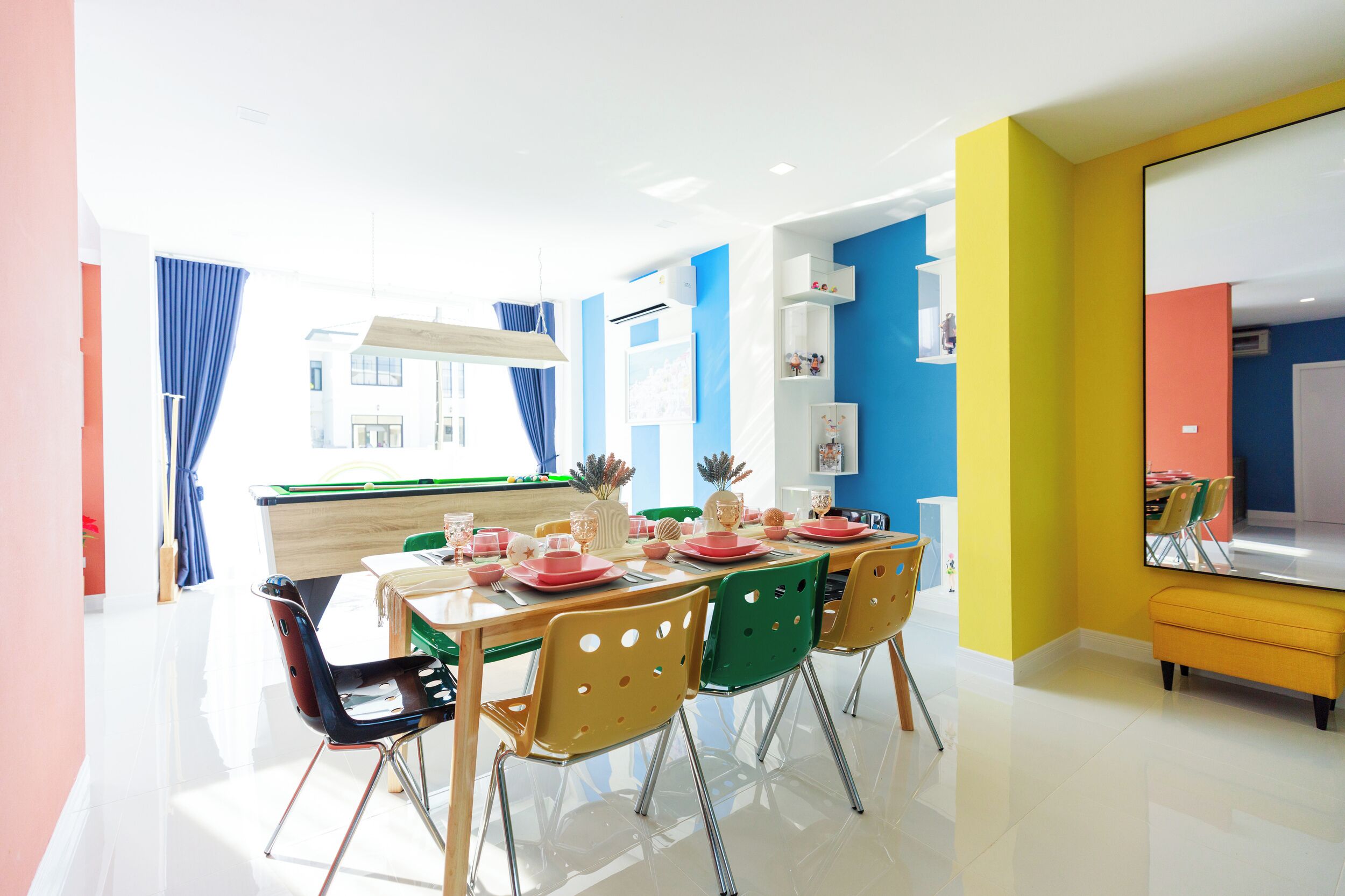 Un comedor luminoso y colorido con una mesa preparada para cuatro, sillas y un espejo grande en la pared.
