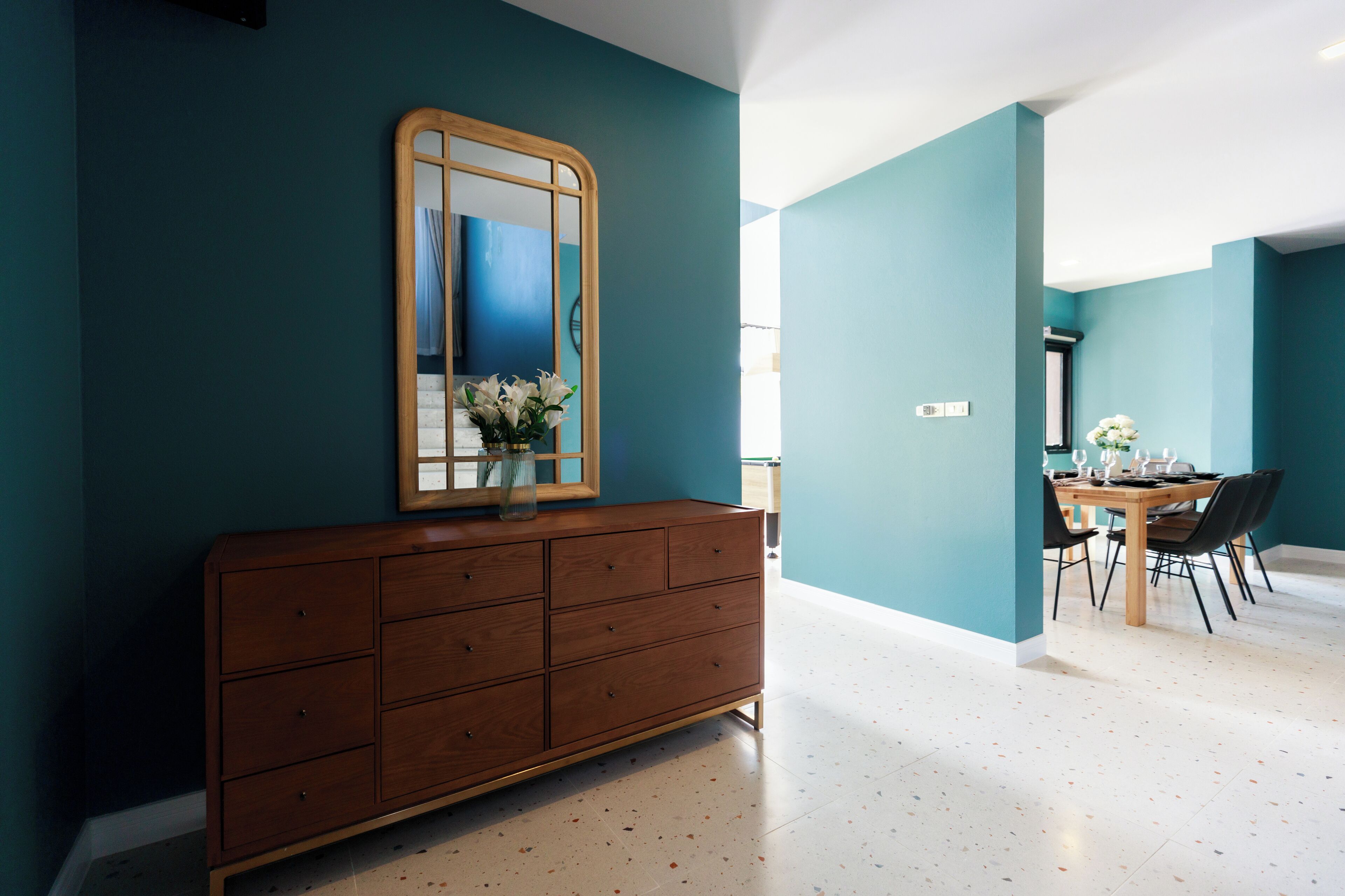 Una habitación con paredes verde azulado, un aparador de madera con espejo, un florero con flores y una zona de comedor con sillas y una mesa.