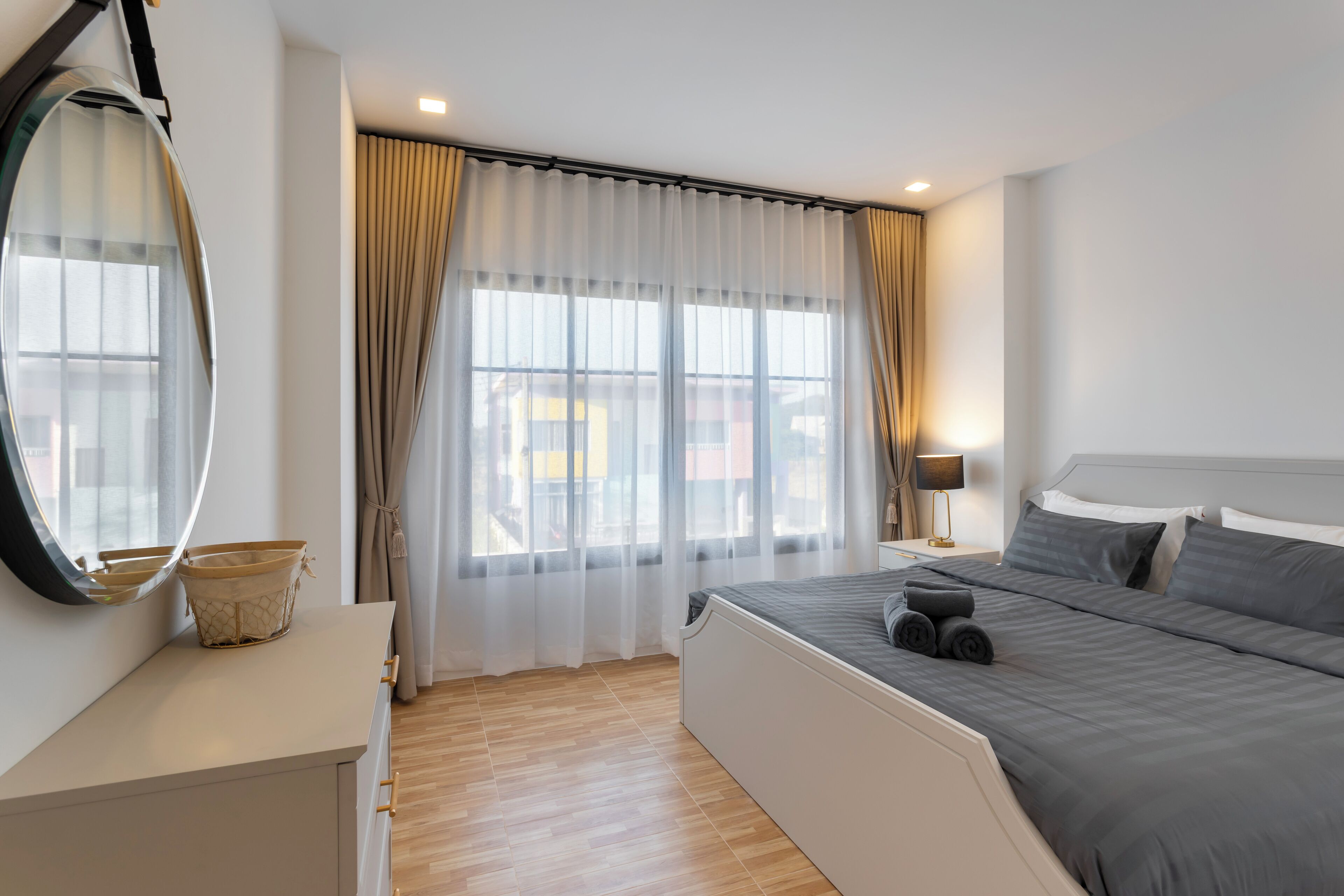 Een slaapkamer met een bed, nachtkastje met lamp, raam met gordijnen en een spiegel.