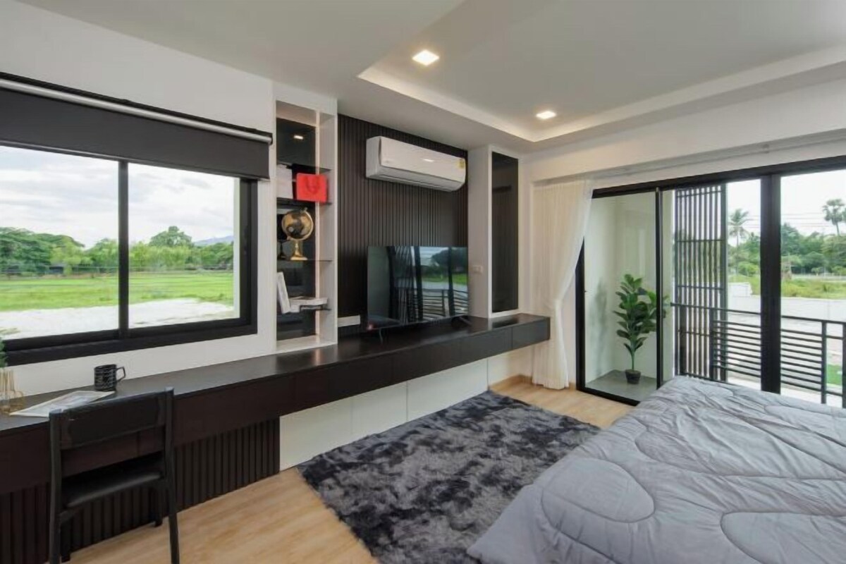A modern bedroom with a large bed, a desk with a computer, and a TV. There is a balcony with a view of greenery.