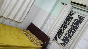 Un dormitorio con cama, mesita de noche, un mueble pequeño, ventana con celosía decorativa y un equipo de aire acondicionado.